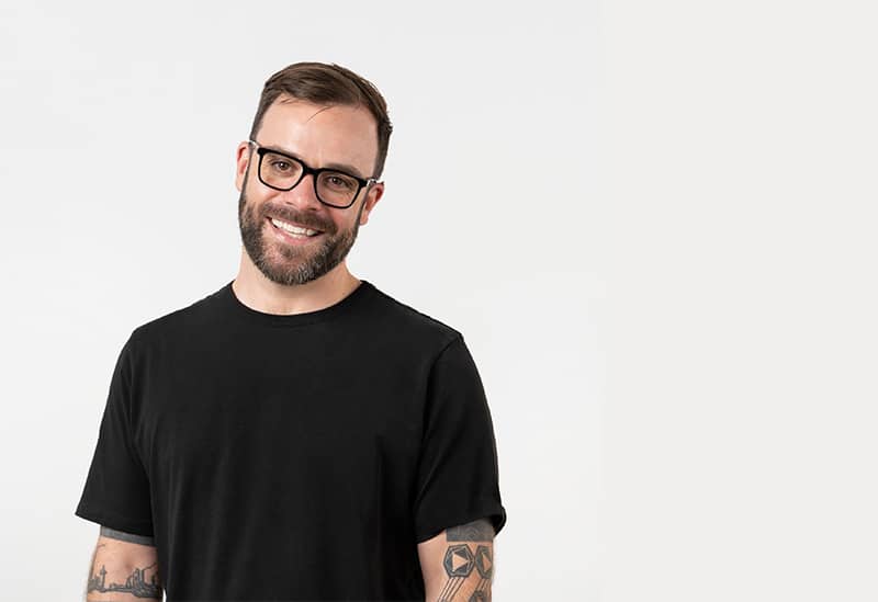 Smiling man with glasses, tattoos, in black shirt.